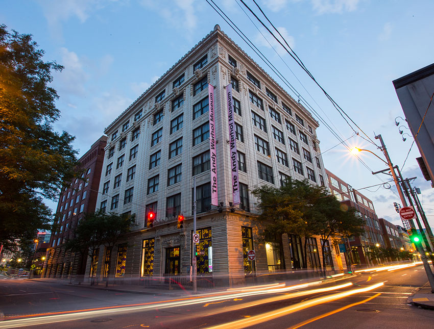 The Andy Warhol Museum, a historic building with banners, illuminated at dusk amid city streets and passing cars.