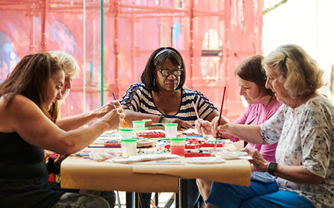 A group of four women gathered around a table, engaged in a painting activity with colorful paints and brushes, in a bright, cheerful setting.