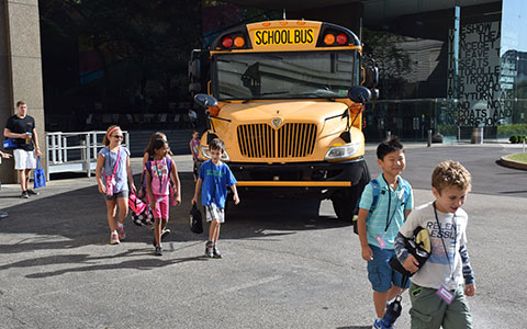 A yellow school bus parked at the museum, as children walk towards it, carrying backpacks and chatting happily.