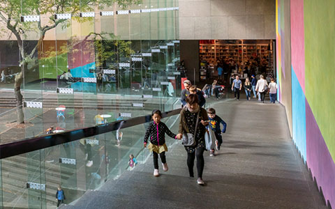 People walking down a modern, colorful corridor with glass walls, leading to a busy public space and bookstore.