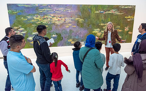A museum guide discusses Monet's water lilies painting with a diverse group of attendees, including children and adults.