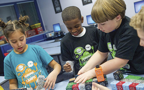 A group of children engaged in a hands-on science activity, building with colorful blocks on a table in a bright classroom setting.