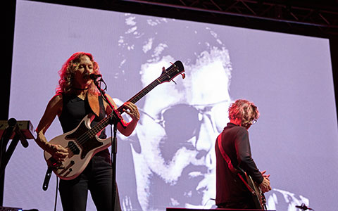 A woman performs energetically on stage with an electric guitar while a guitarist plays behind her, against a large projected backdrop.