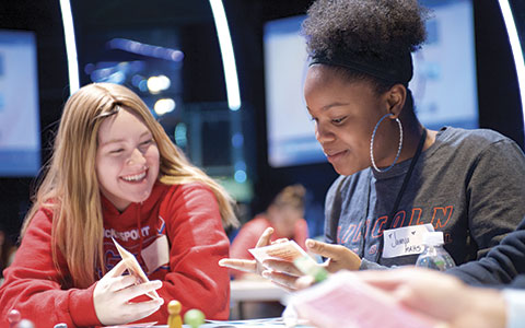 Two teenagers engage in a game, focusing on cards and strategy, surrounded by a vibrant, colorful gaming environment.