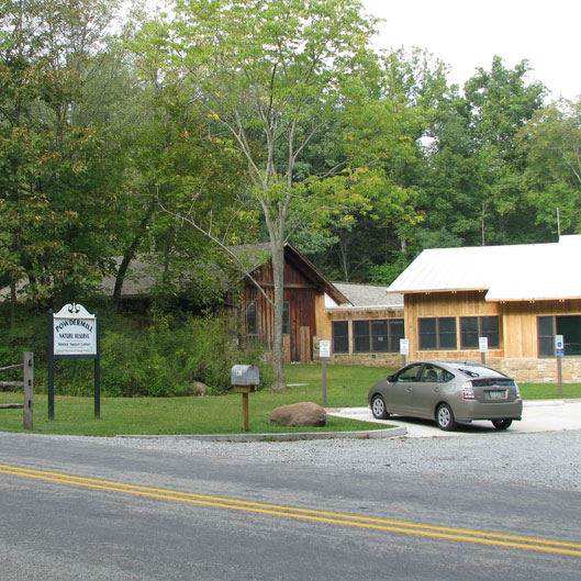 A gravel parking area with a gray car, flanked by rustic buildings and surrounded by greenery at the Powder Mill Nature Reserve.