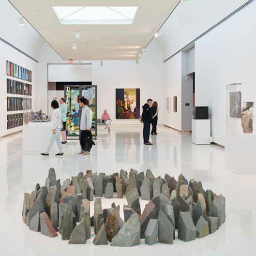 A modern art gallery scene featuring visitors exploring various artworks, with a circular stone installation in the center.