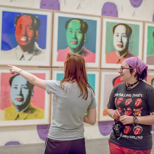 Two women observe colorful framed portraits in an art gallery, one pointing at the artwork while the other listens attentively.
