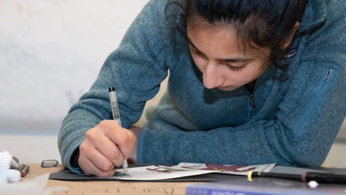 A person wearing a blue sweater writes with a pen on paper, surrounded by art supplies on a wooden table.