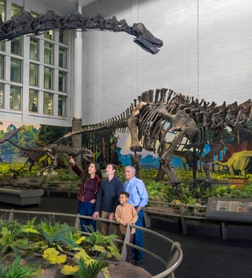 A family admires dinosaur fossils in a museum exhibit surrounded by colorful murals and lush plant displays.