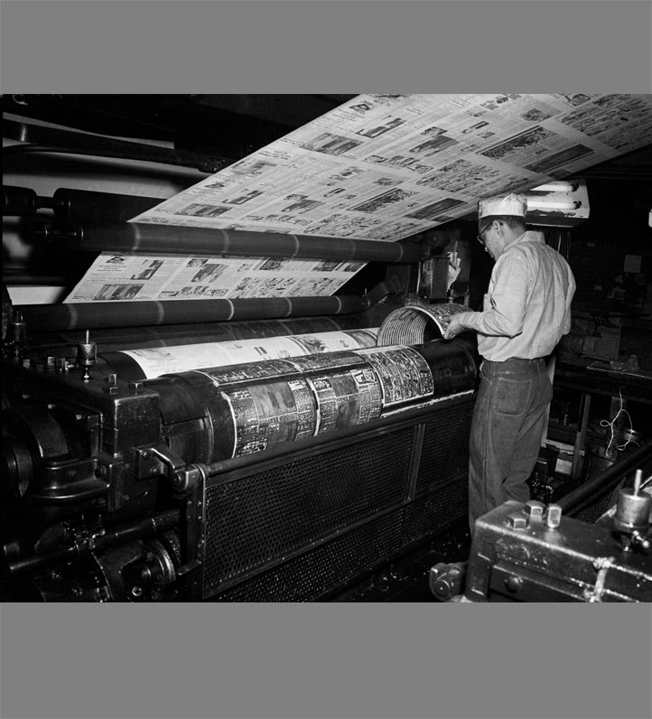 A man operates a vintage printing press, overseeing newspaper production as printed sheets roll out above him in a dimly lit factory.