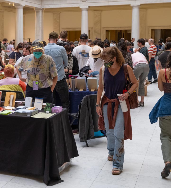 A crowded indoor event filled with people browsing at tables displaying various books, with a woman walking past.