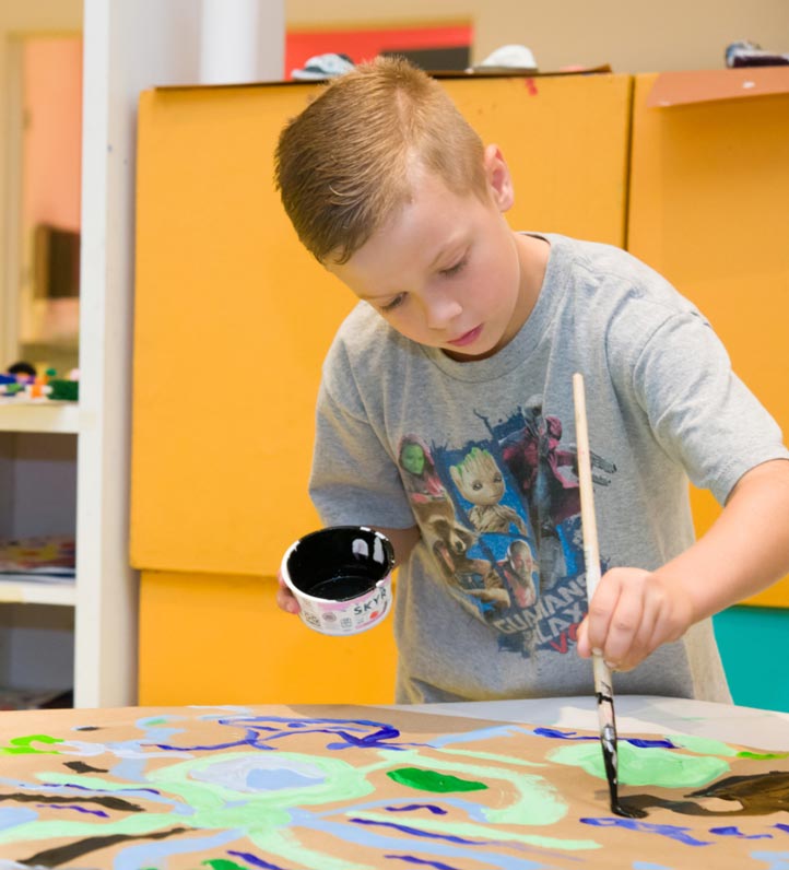 A child paints a colorful abstract design on a large canvas, holding a black paint container in one hand and a brush in the other.