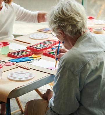 A woman with gray hair paints at a table, surrounded by art supplies, as another person works nearby.