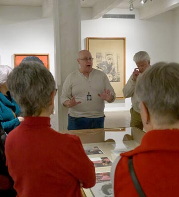 A group of visitors listens attentively to a museum guide discussing artwork in a gallery setting, surrounded by framed pieces.