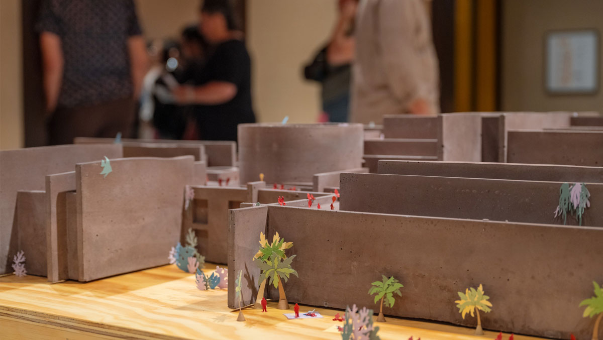 A detailed architectural model featuring gray concrete structures and colorful miniature trees, with people observing in the background.