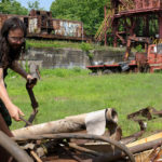 Two men sorting through rusted scrap metal in front of a rusted abandoned steel mill.