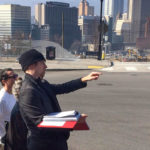 Group of people standing together with the city of Pittsburgh behind them. One man is pointing and holding a book.