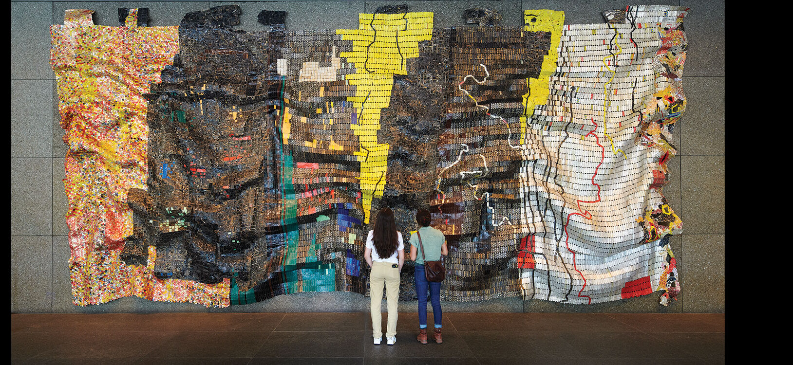Two people standing in front of a large tapestry made of bottle caps and metal scraps.