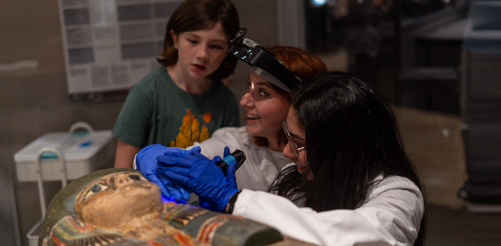 Two museum conservators working on a colorful egyptian sarcophagus, with a young girl standing near them watching.