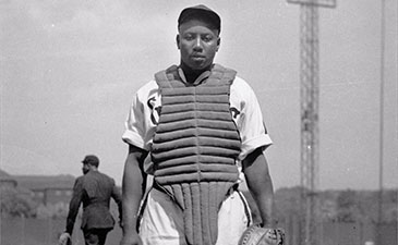 A black and white photo of an Black baseball player wearing a catchers vest.