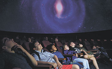 A group of children seated in a planetarium, staring at a star filled screen.