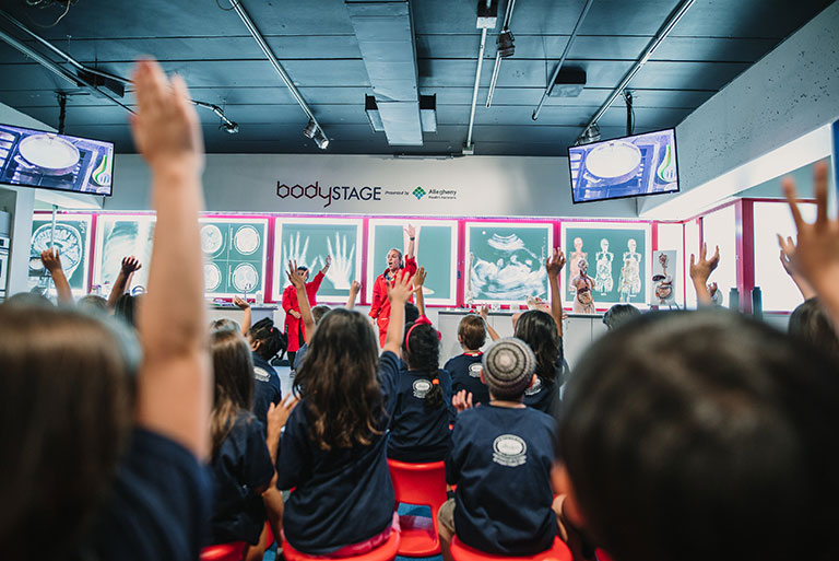 Two museum educators in red lab coats on a stage with young students in the foreground raising their hands.