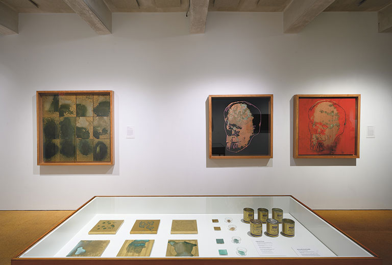 An installation view of an art exhiti, with 3 paintings on a wall behind a table with smaller objects.