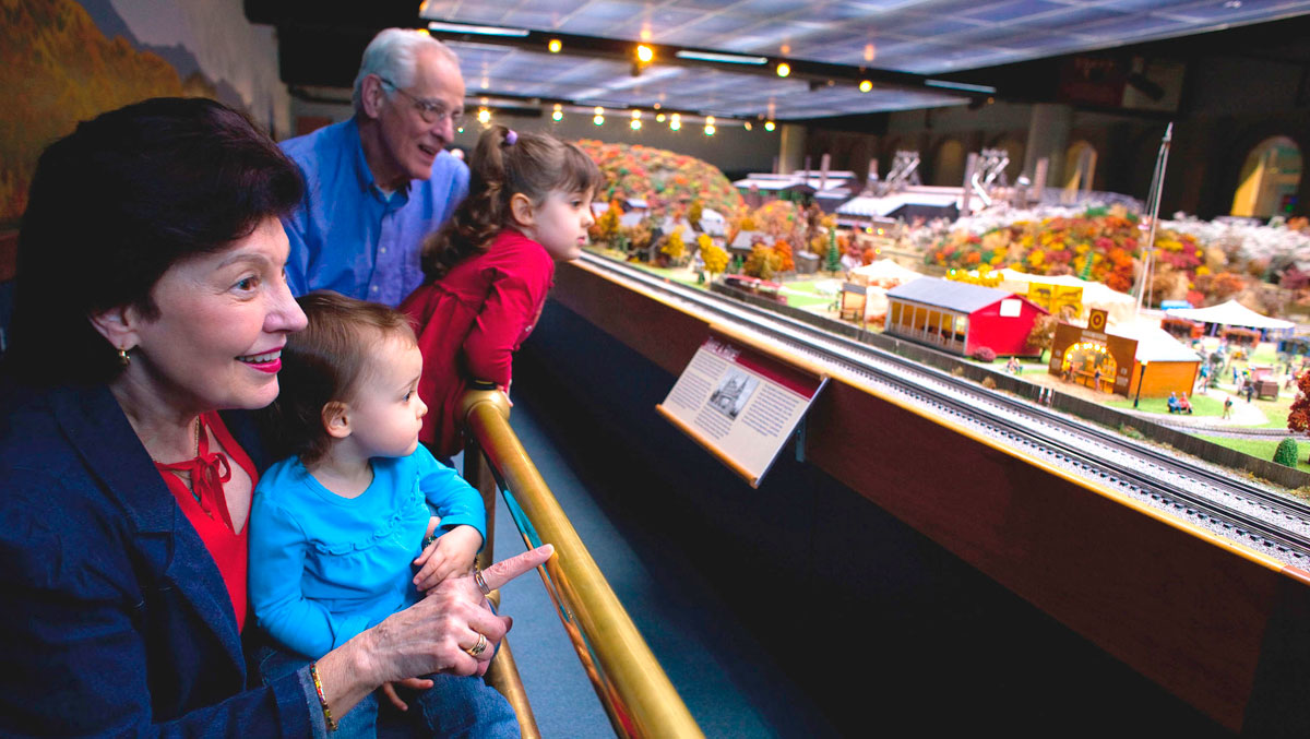 A family observes a detailed miniature train layout, with colorful trees and small buildings.