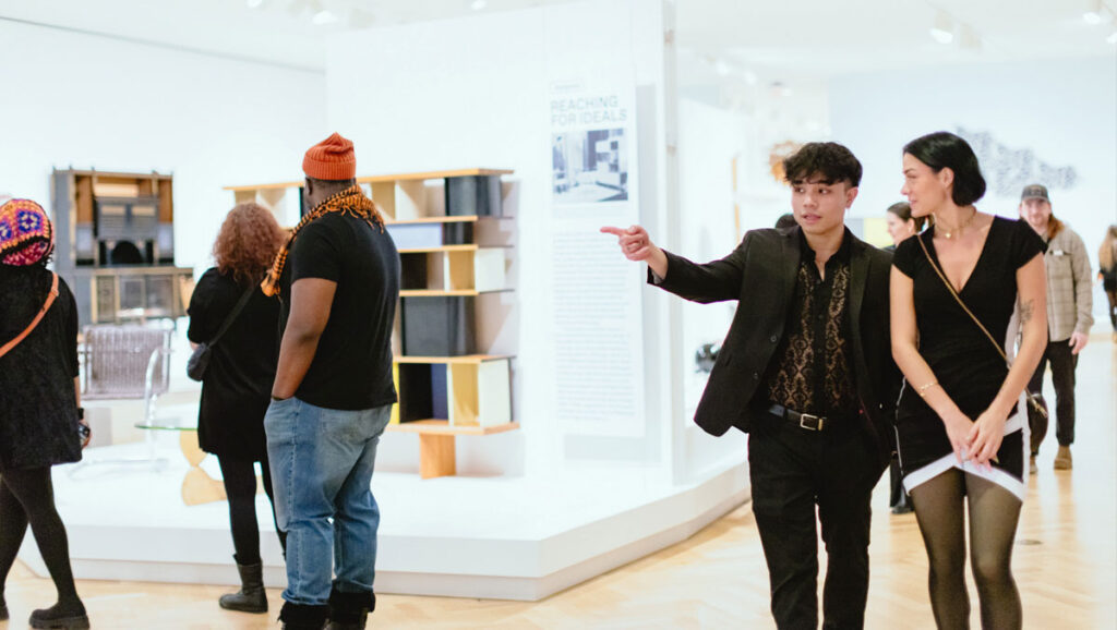 Several people explore a modern exhibit filled with stylized furniture and informative signs in a bright, minimalist gallery.