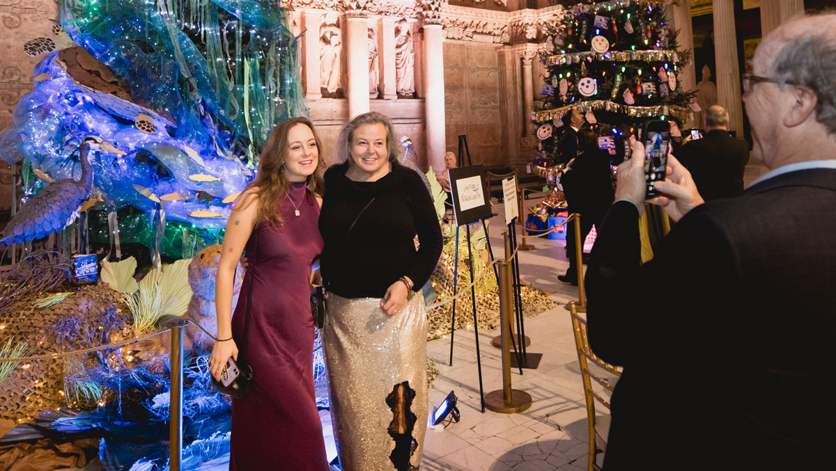 Two women pose together against a vibrant holiday backdrop featuring an elaborate decoration with lights and a festive tree.