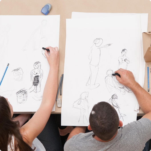 Two artists sketch figures on large sheets of paper in a shared workspace, surrounded by drawing tools and materials.