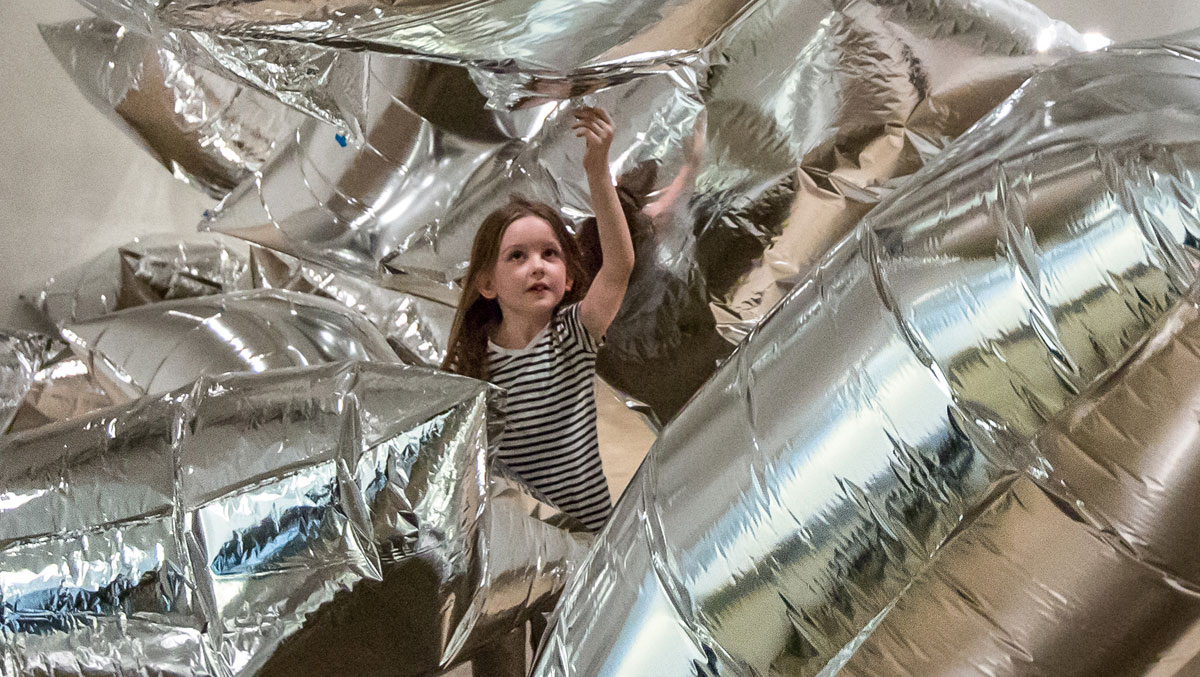 A child in a striped shirt plays among large, shiny silver inflatable shapes in a bright indoor space.