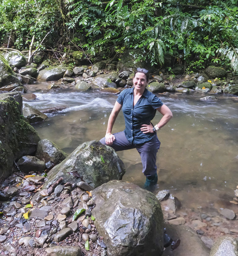 A person stands confidently on a rock by a flowing stream, surrounded by lush green foliage in a tropical forest setting.