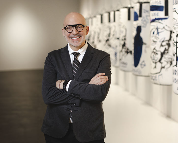 A man in a suit stands confidently with arms crossed in an art gallery, featuring colorful artworks on white elongated panels.