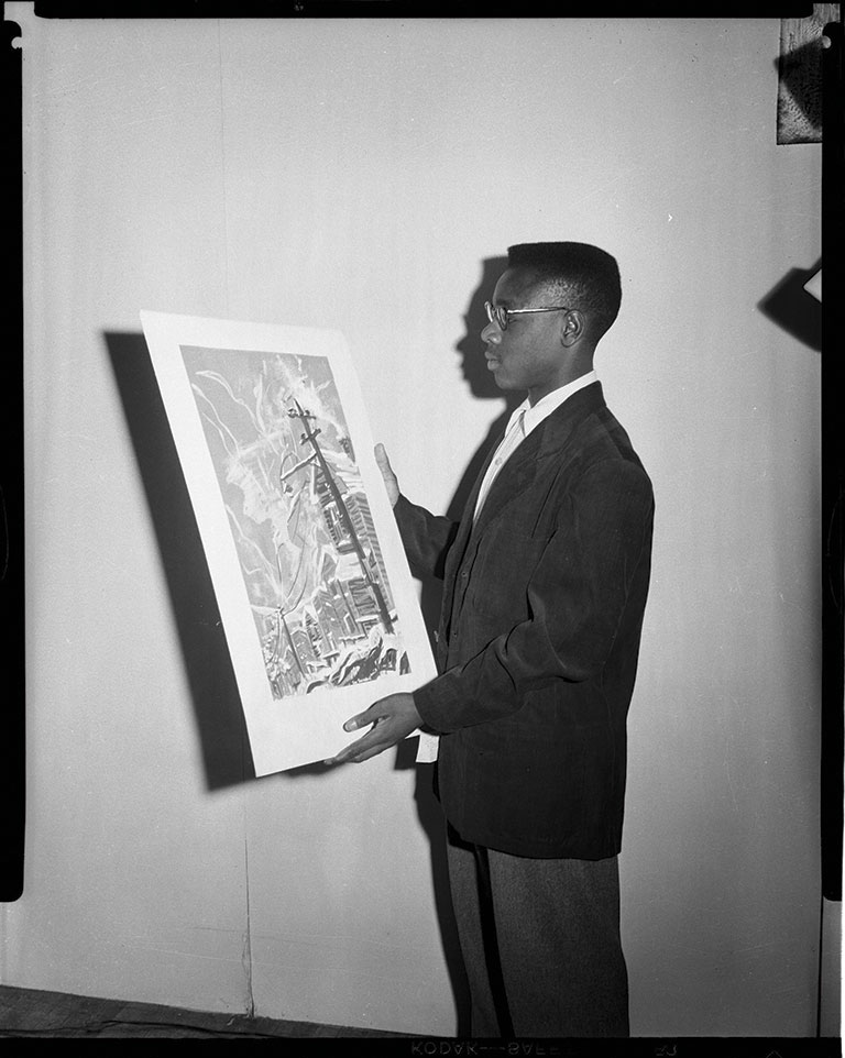 A young man in a suit holds a large, colorful artwork depicting a cityscape against a plain background.