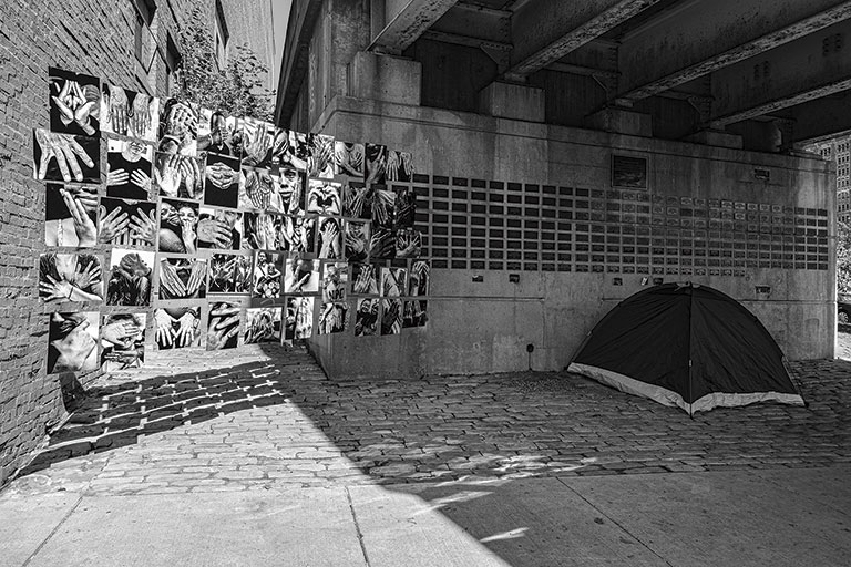 A black and white image of a tent under a bridge, with a wall of hand photos displayed next to it, evoking themes of shelter and creativity.