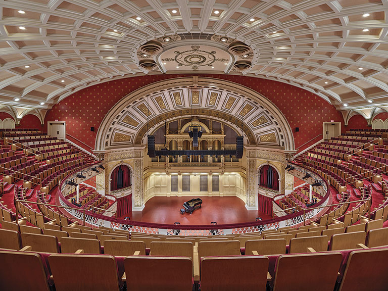 A grand concert hall interior featuring ornate architecture, red accents, and a grand piano on stage, with tiered seating visible.