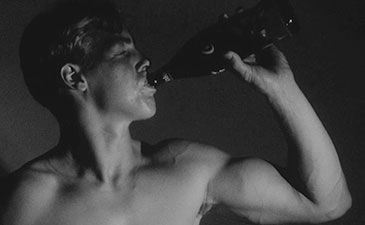 A still from a black and white Warhol movie of a bare chested young man, drinking out of a bottle