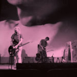 Black and white photo of a band on stage playing music with a film of ttwo people kissing projected behind them