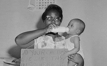 A vintage photo of a woman holding a sign and feeding a bottle to her baby.