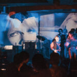 Color photograph of three of the members of the Velvet Underground performing on a stage as images are being projected onto the walls behind them.