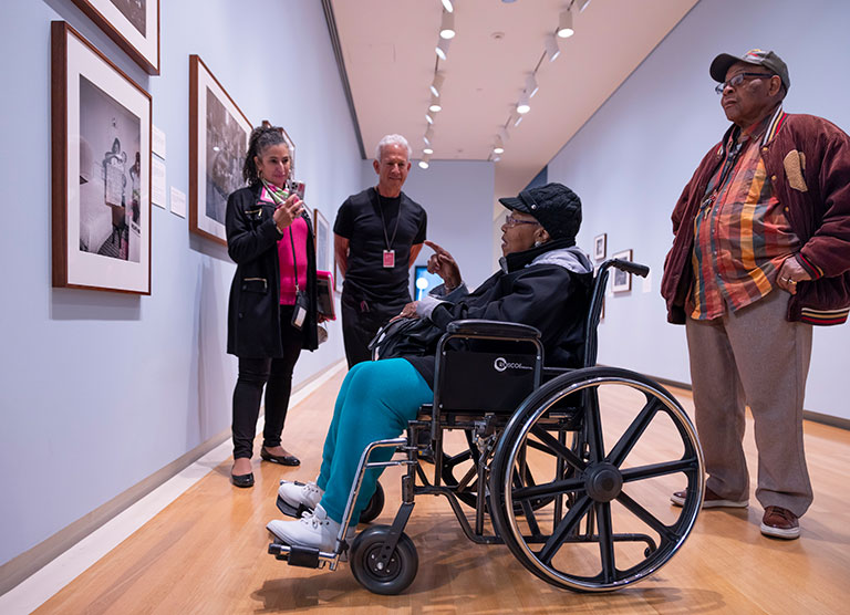 An elderly woman in a wheelchair in an art gallery looking at a photo as three other people look on.