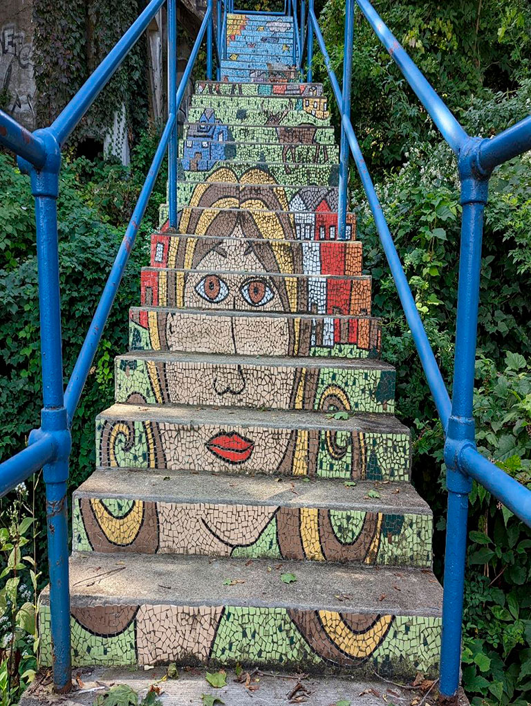 A colorful, mosaic-tiled staircase featuring a woman's face and houses, surrounded by greenery and blue handrails.