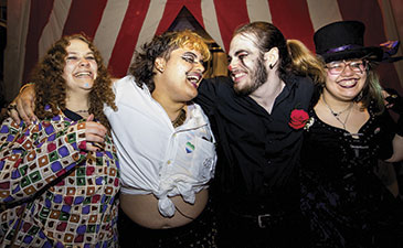 Four individuals pose together in a festive environment, dressed in colorful and eclectic outfits, showcasing a lively celebration.