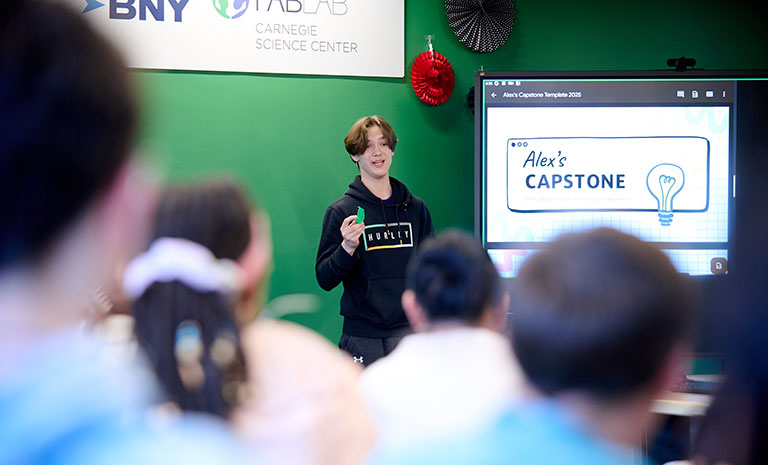 A presenter stands in front of a screen displaying "Alex's Capstone," while an audience listens attentively in a classroom setting.
