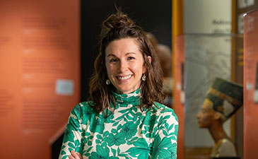 A person in a green floral top stands with arms crossed in a museum, with colorful exhibit walls and artifacts displayed nearby.