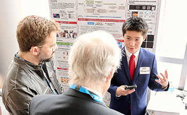 A student presenter in a suit gestures while discussing research with two attendees in front of an academic poster display.