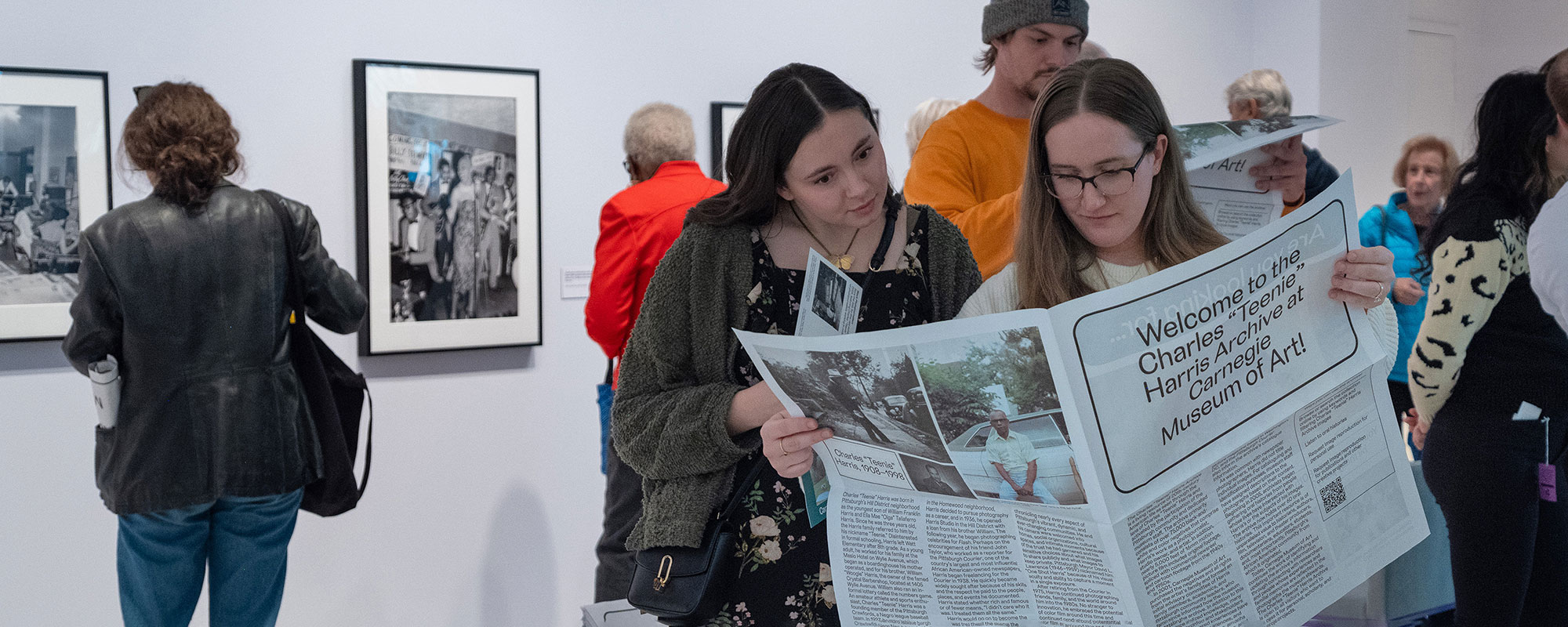 Two young women examine a large newspaper in an art gallery, while other visitors observe black-and-white photographs on the walls.