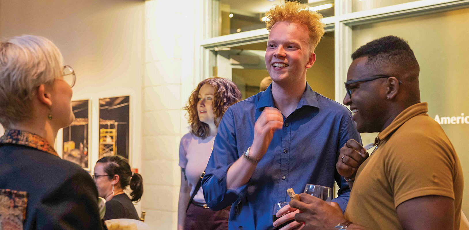A group of three people engage in conversation at a social event, smiling and laughing, with others in the background.
