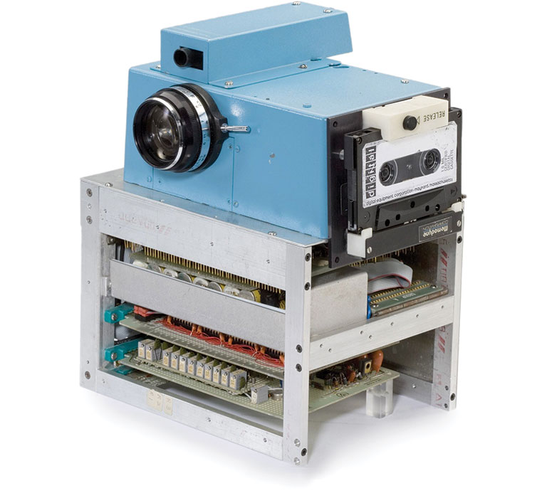 A vintage blue camera-like device with a large lens, attached to a cassette tape, and a visible circuit board structure below.